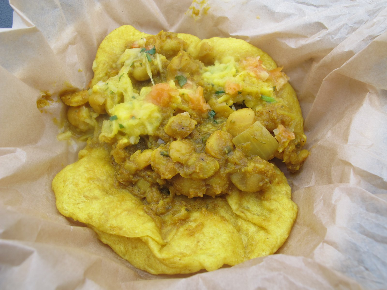 Close-up of doubles on brown paper, with chickpeas and colourful chutneys spread over two overlapping pieces of golden fried bara.
