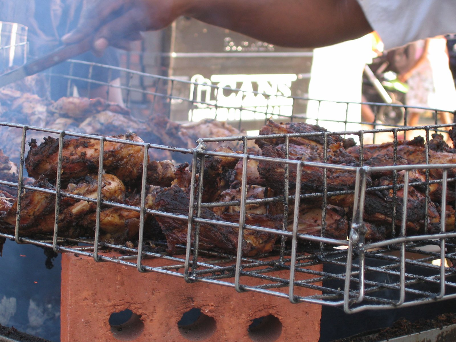 Pieces of browned jerk chicken in a wire barbecue rack, with smoke rising as a cook tends the grill.