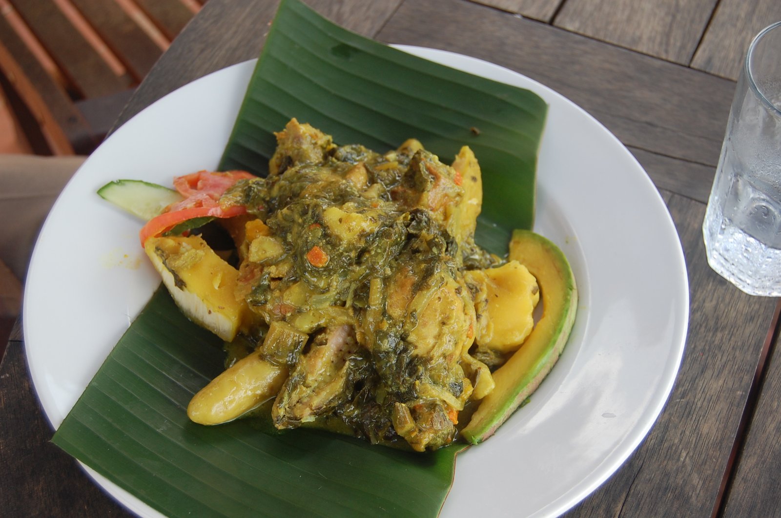 A plate of oil down with yellow breadfruit pieces, chicken and cooked greens on a banana leaf, beside sliced avocado, tomato and cucumber.