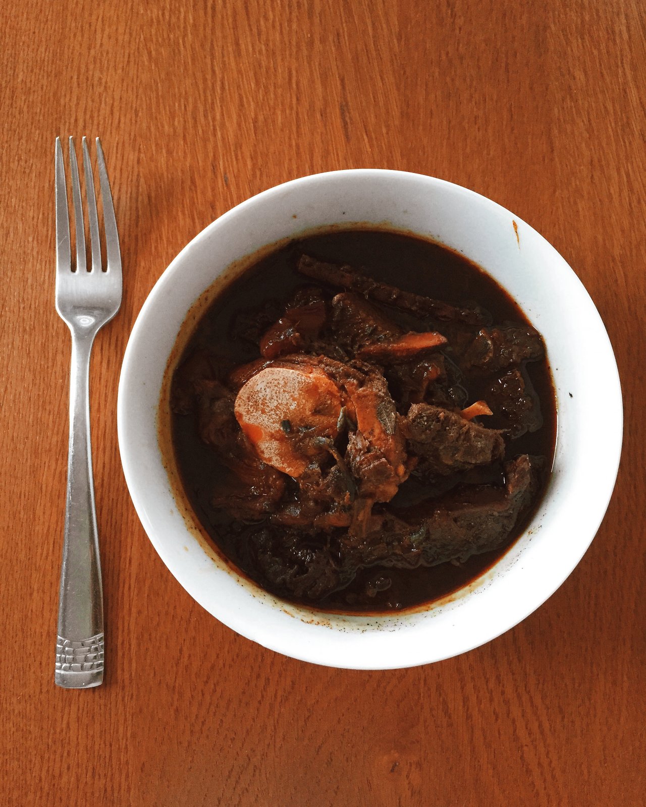 A white bowl of Guyanese pepperpot with bone-in meat in a dark sauce on a wooden table.